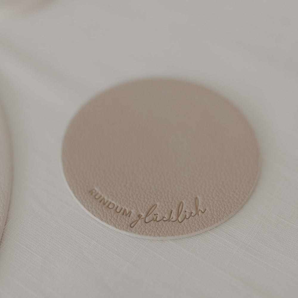 Untersetzer in Taupe mit Prägung „rundum glücklich“ auf hellem, schlicht gedecktem Tisch.