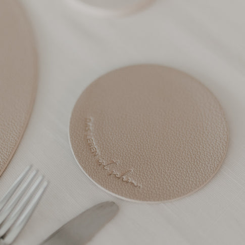 Beiger Untersetzer mit Schriftzug „Das Leben ist schön“ auf weiß gedecktem Tisch.