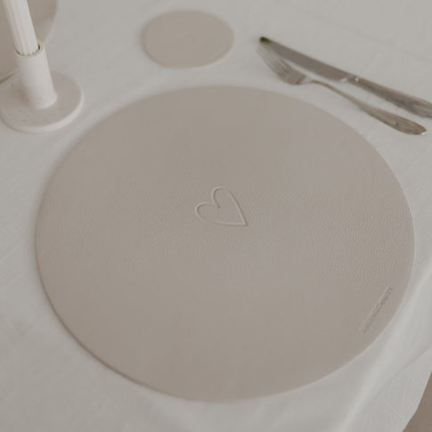 Rundes Platzset mit Herzprägung in Creme, dekoriert auf weißer Tischdecke mit Besteck.