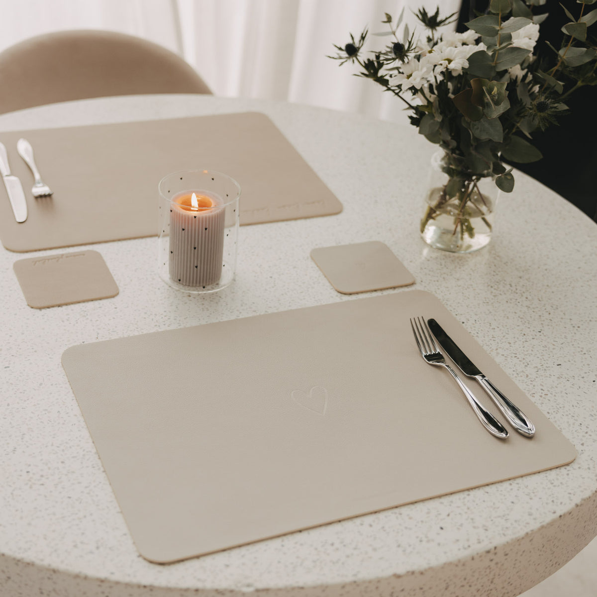 Gedeckter Tisch mit cremefarbenen Platzsets und quadratischen Untersetzern, Kerze und Blumenstrauß