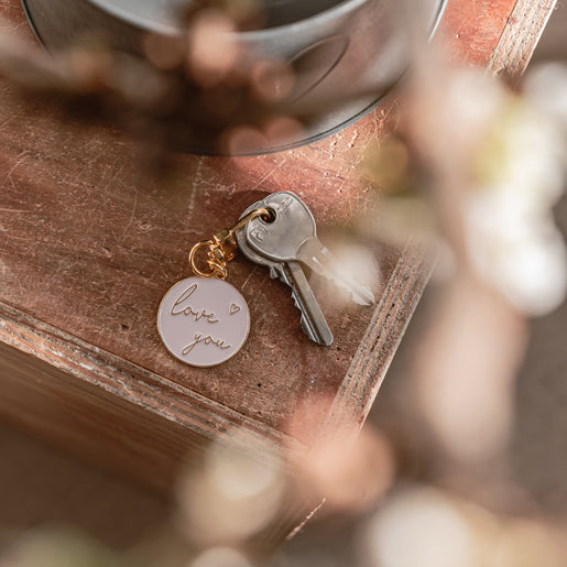 Ein Schlüsselanhänger in der Farbe rosa & gold mit der Aufschrift *love you* liegt auf einem Holztisch.