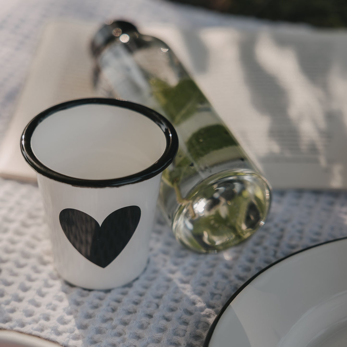 Weißer Emaille-Becher mit großem schwarzem Herz neben Trinkflasche auf einer Picknickdecke.