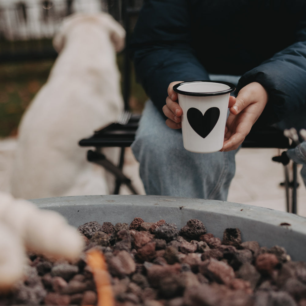 Person hält Emaille Tasse Herz am Lagerfeuer – ideal für warme Getränke an kühlen Abenden.