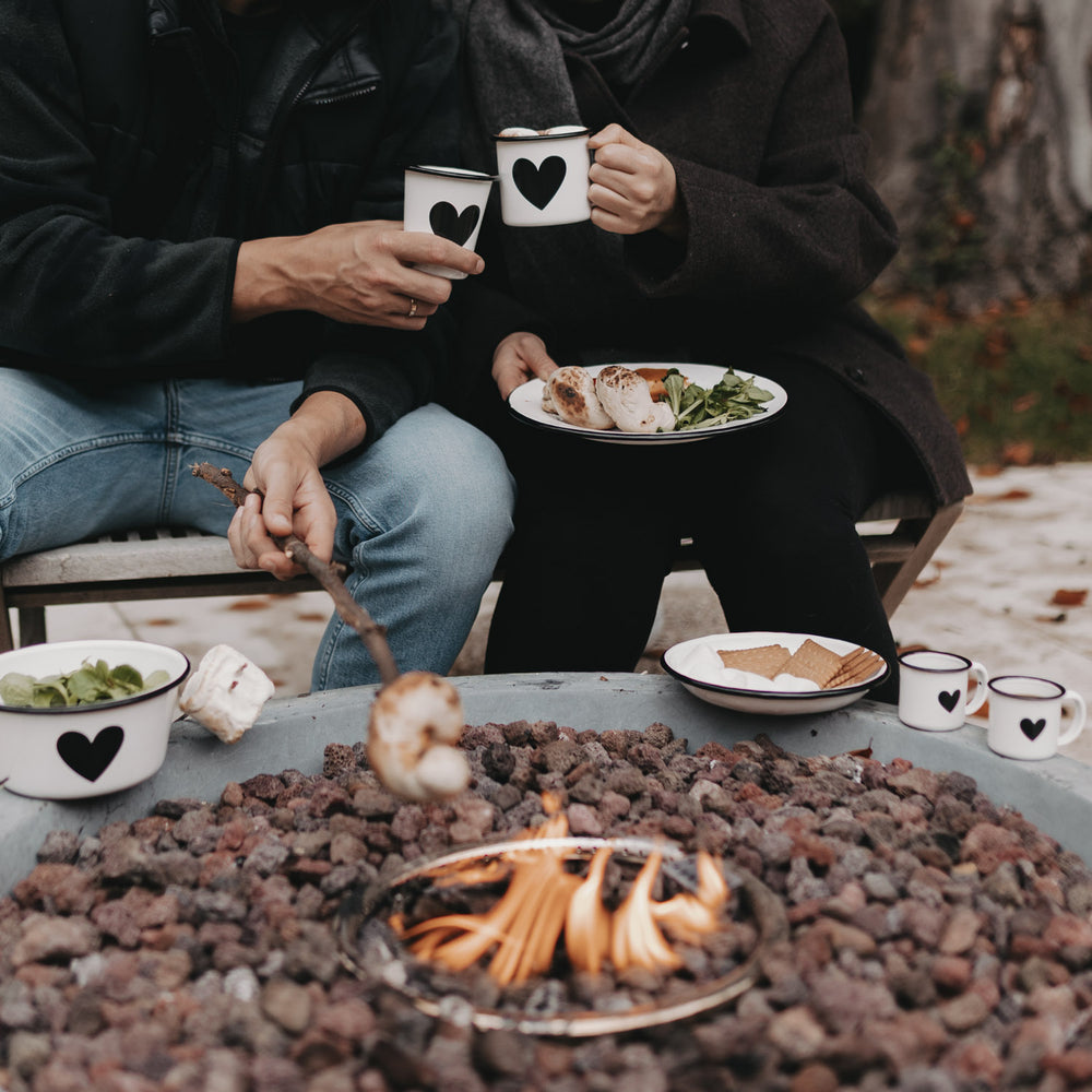 Zwei Menschen stoßen mit Emaille Tassen Herz am Lagerfeuer an – gemütliches Beisammensein im Freien.