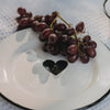Weißer Emaille-Speiseteller mit schwarzem Herz, belegt mit einer Weintraube, im sommerlichen Picknick-Setting.