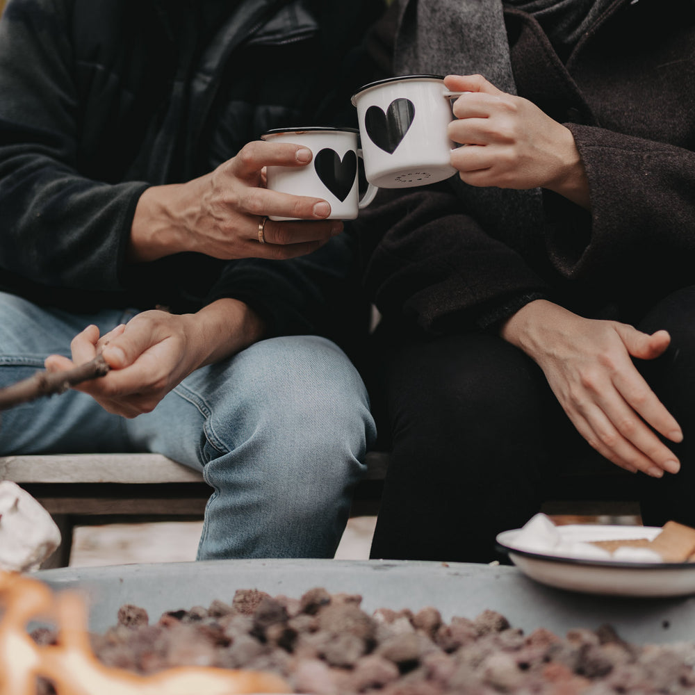 Zwei Personen stoßen mit Emaille Tassen Herz am Lagerfeuer an – gemütlicher Moment im Freien.