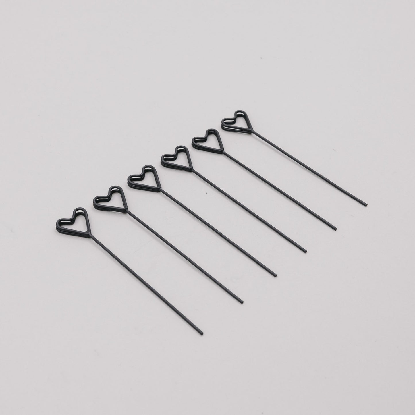 Sechs schwarze Herz-Stecker aus Draht nebeneinander auf hellem Untergrund arrangiert.