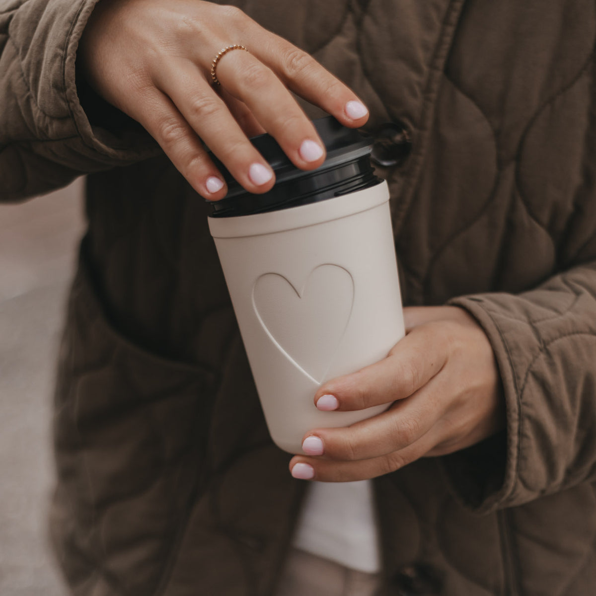 Cremefarbener To-Go-Becher mit Herzmotiv und schwarzem Deckel wird von einer Frau verschlossen