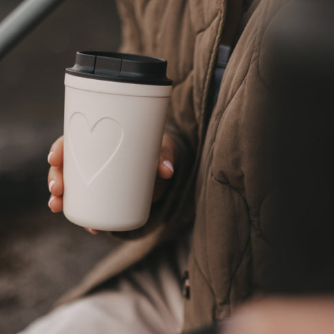 Cremefarbener To-Go-Becher mit Herzmotiv und schwarzem Deckel in der Hand einer Frau