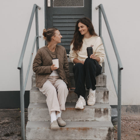 Zwei Frauen sitzen mit schwarzen und cremefarbenen To-Go-Bechern mit Herzmotiv auf einer Treppe