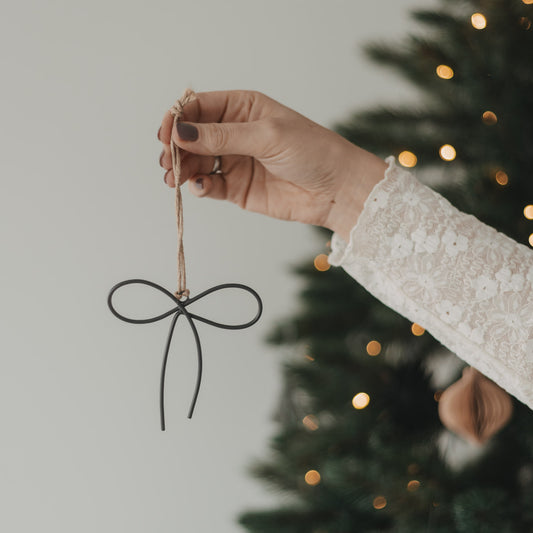 Metallanhänger Schleife in Anthrazit mit Juteschnur als Weihnachtsbaumschmuck vor einem beleuchteten Tannenbaum.