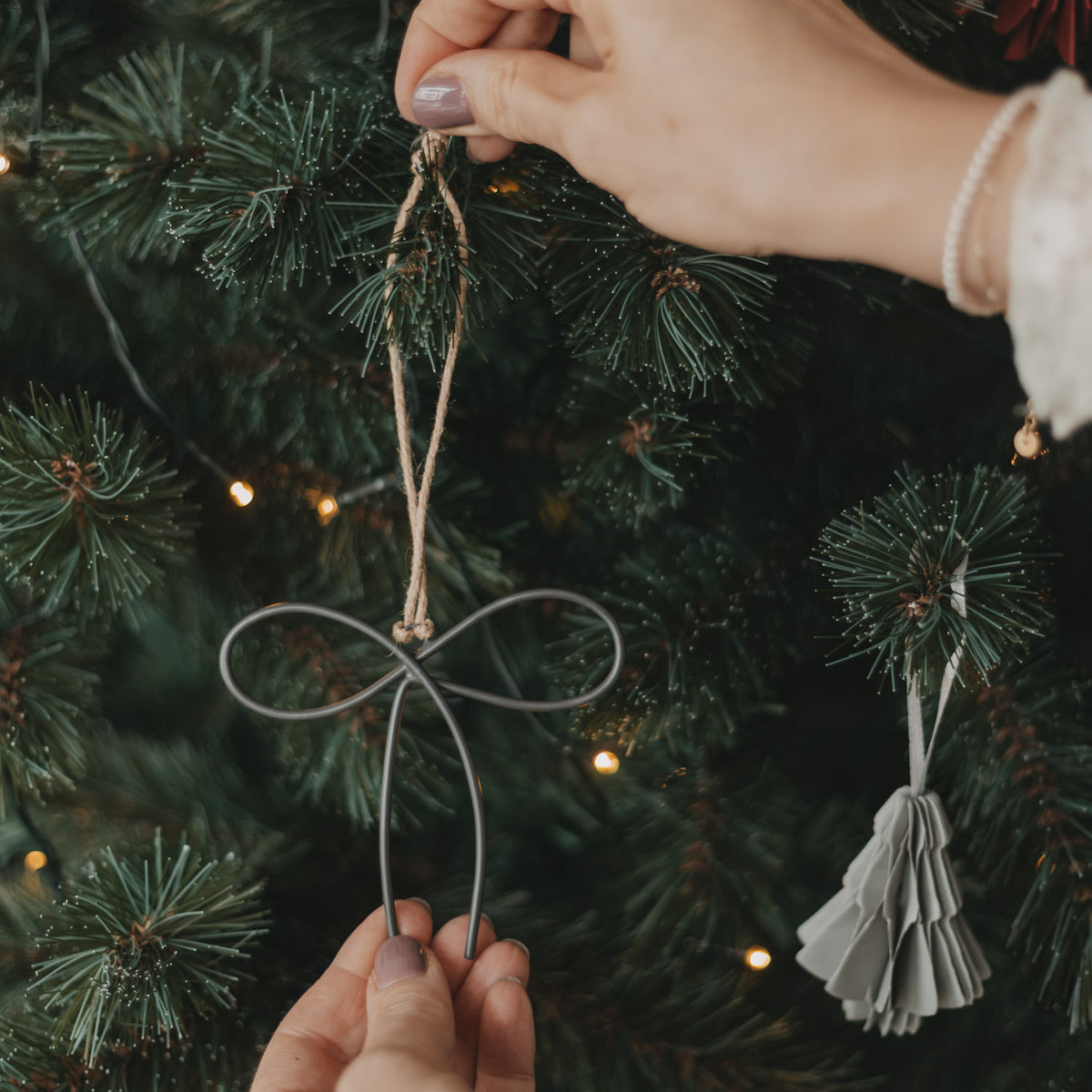 Anthrazitfarbener Metallanhänger in Schleifenform wird mit Juteschnur als Christbaumschmuck am Weihnachtsbaum befestigt.