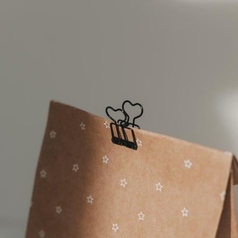 Schwarzer Herz-Clip verschließt eine braune Geschenktüte aus Kraftpapier mit weißen Sternen.