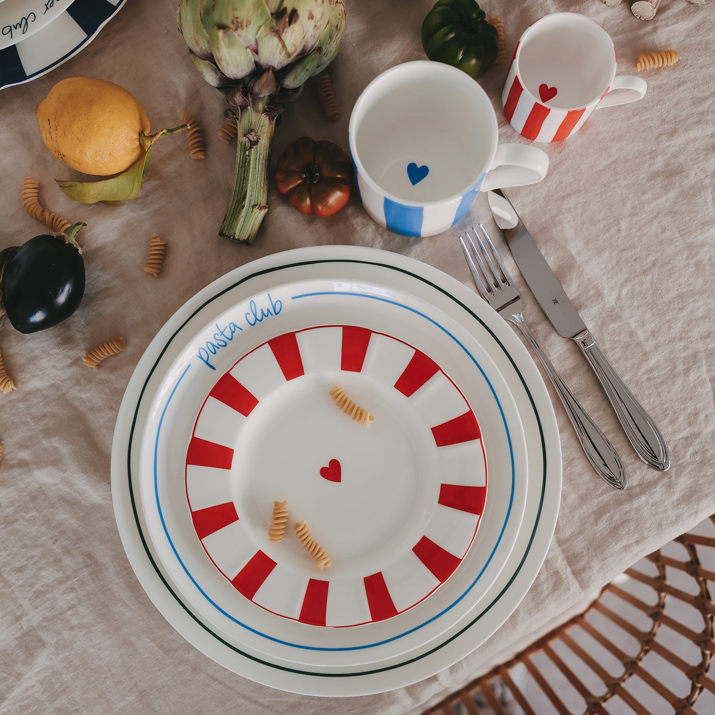 Arrangement aus drei verschiedenen Tellern: roter, welliger Frühstücksteller mit Herz, hellblauer „Pasta Club“ Speiseteller und grüner Platzteller mit Herz, dekoriert mit Pasta, Gemüse, Tassen und Besteck auf hellem Leinentisch.