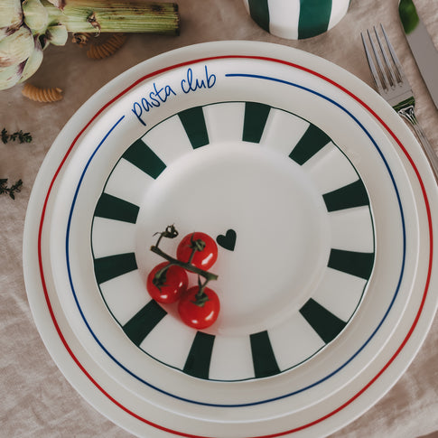 Speiseteller „pasta club“ mit blauem Rand gestapelt mit 2 weiteren bunten Porzellantellern, dekoriert mit Cherrytomaten auf hellem Leinentisch.