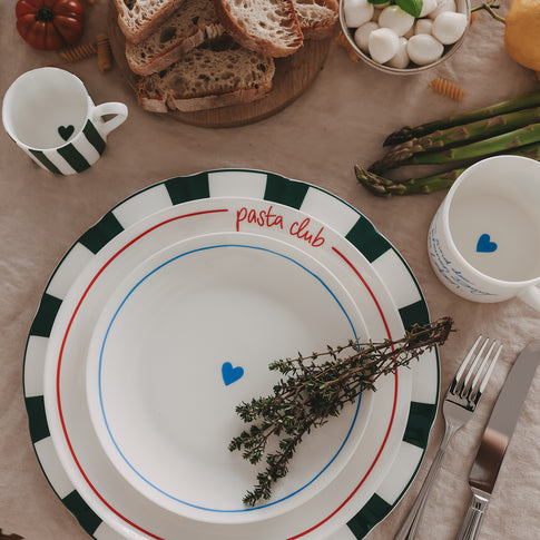 Gestapeltes Porzellan mit grünem Streifenrand, roter "Pasta-Club"-Schrift und blauen Herzdetails, kombiniert mit dazu passenden Tassen, Brot, Mozzarella und Spargel auf natürlicher Leinentischdecke.