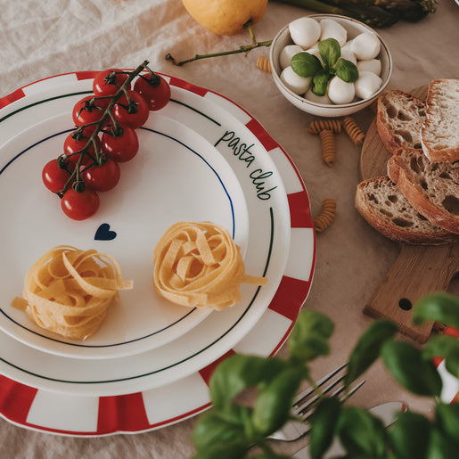Gestapelte Porzellanteller, dekoriert mit frischer Pasta, Tomaten und Brot auf Leinentischdecke.
