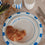 Croissant auf weißem „breakfast club“-Teller mit blauem Rand, umgeben von blau-weiß gestreiften Tassen, Tellern, Besteck und verstreuten Nudeln sowie grünem Spargel auf Leinentisch.