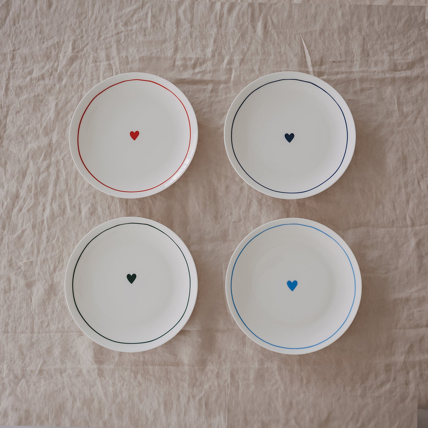 Vier runde Porzellanteller mit farbigen Rändern und kleinem Herz in der Mitte in verschiedenen Farben auf Leinentuch.