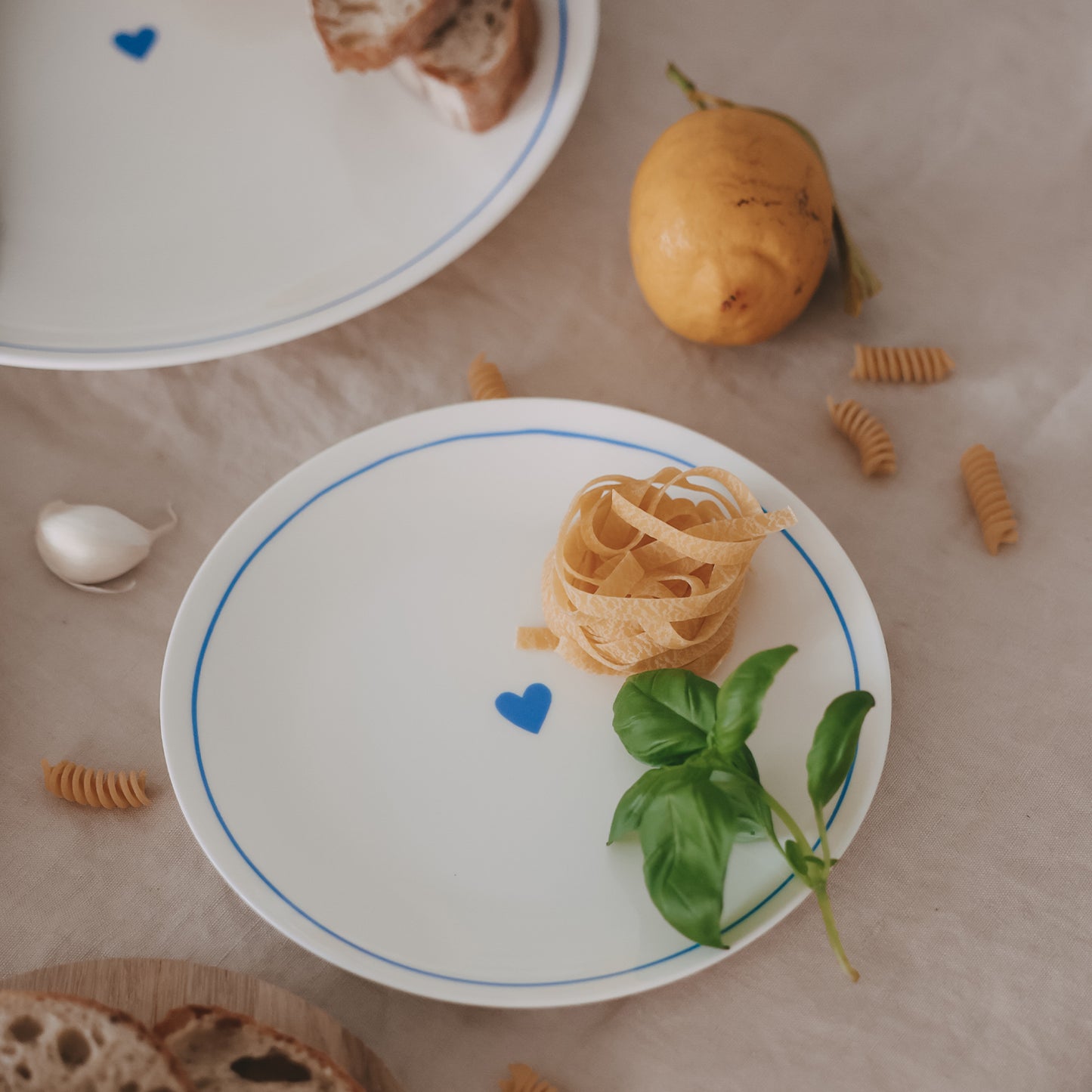 Runder Porzellanteller mit hellblauem Rand und Herzdetail, dekoriert mit Pasta und Basilikum auf Leinentuch.
