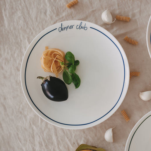 Weißer „dinner club“-Teller mit dunkelblauem Rand, belegt mit Tagliatelle, Aubergine und frischem Basilikum, umgeben von Knoblauch und Pasta auf Leinentisch.