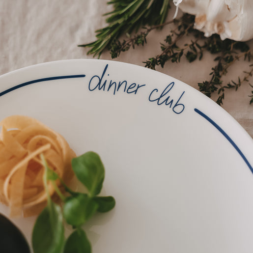 Detailansicht eines weißen „dinner club“-Tellers mit dunkelblauer Schrift und Rand, garniert mit Pasta und frischen Kräutern.