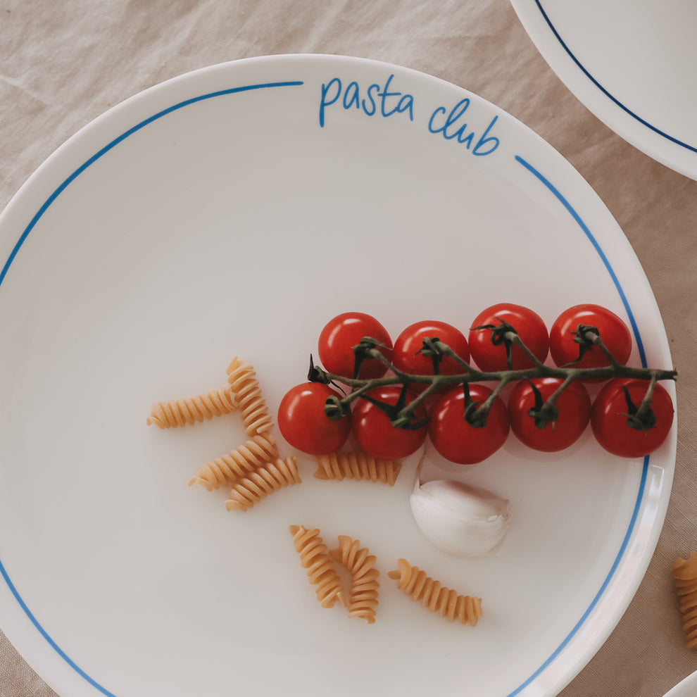 Speiseteller „pasta club“ mit hellblauem Rand, dekoriert mit Cherrytomaten, Fusilli und Knoblauch auf hellem Leinentisch.