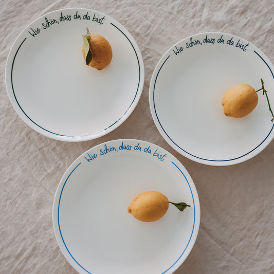 Drei Platzteller „Wie schön, dass du da bist“ in dunkelblau, grün und hellblau, jeweils mit Zitrone auf hellem Leinentisch arrangiert.