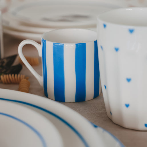 Gestreifte kleine Tasse in Hellblau neben einem Becher mit kleinen blauen Herzmotiven, arrangiert auf gedecktem Tisch mit Teller und Pasta im Hintergrund.