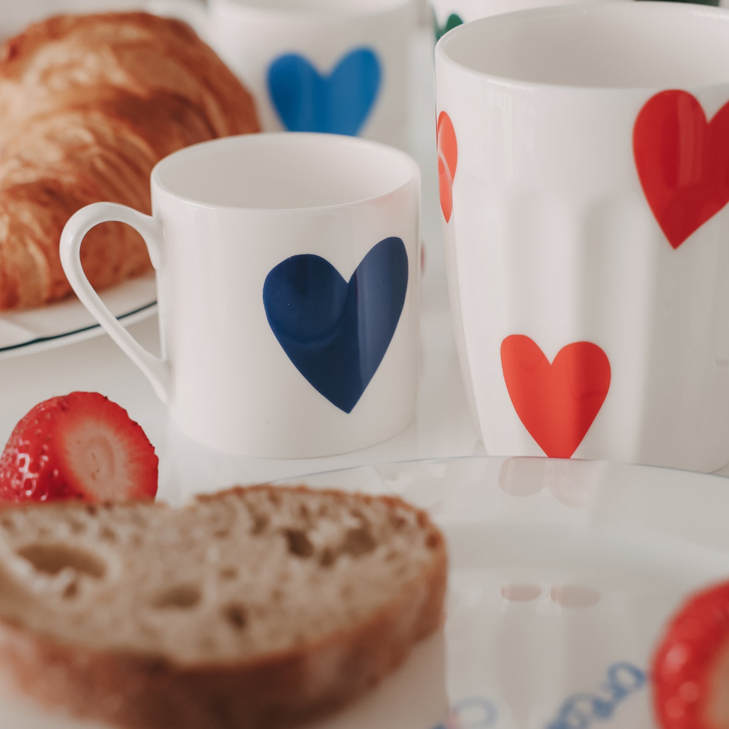 Frühstücksszene mit kleiner Tasse (dunkelblaues Herz) neben Croissant und Erdbeeren, weitere Tassen im Hintergrund.