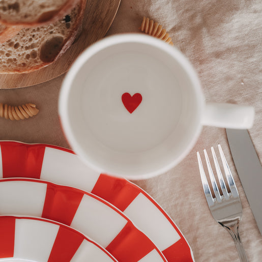 Draufsicht in Tasse mit rotem Herz, umgeben von rot gestreiftem Geschirr und Besteck auf Leinentischdecke