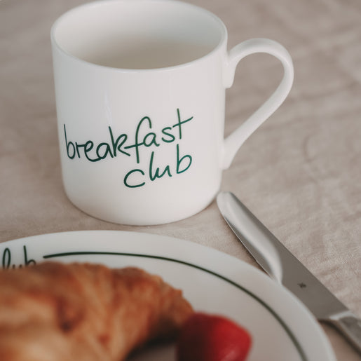Weiße Tasse mit grünem „breakfast club“-Schriftzug neben Croissant und Messer auf gedecktem Tisch