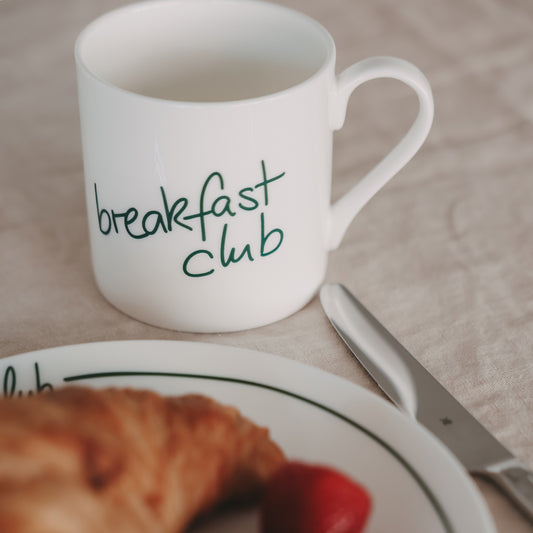 Weiße Tasse mit grünem „breakfast club“-Schriftzug neben Croissant und Messer auf gedecktem Tisch