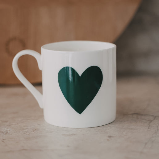 Weiße Tasse mit großem grünem Herz auf der Vorderseite vor warmem Hintergrund