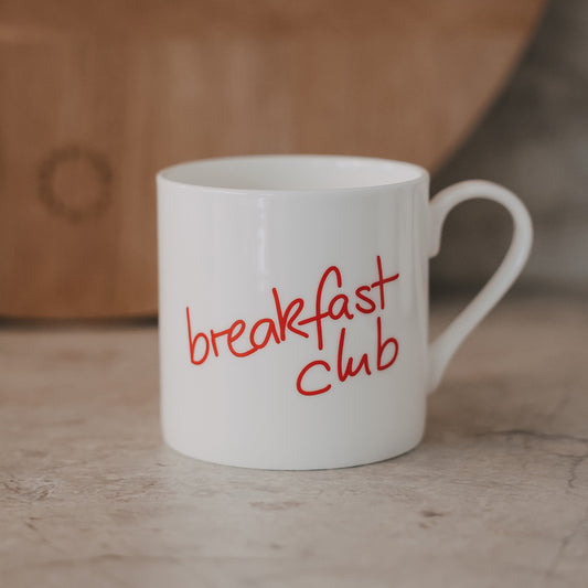 Weiße Tasse mit rotem Schriftzug „breakfast club“ vor warmem Hintergrund