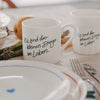 1 weiße Tasse und 1 weißer Becher mit grünem Schriftzug „Es sind die kleinen Dinge im Leben“ auf gedecktem Tisch mit Teller, Besteck und Spargel