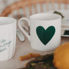 Weiße Tasse mit großem grünem Herzmotiv auf einem gedeckten Tisch, umgeben von Besteck, Pasta und Gemüse