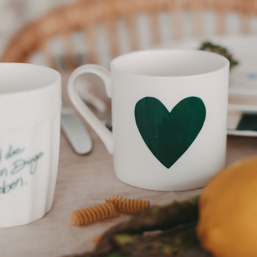 Weiße Tasse mit großem grünem Herzmotiv auf einem gedeckten Tisch, umgeben von Besteck, Pasta und Gemüse