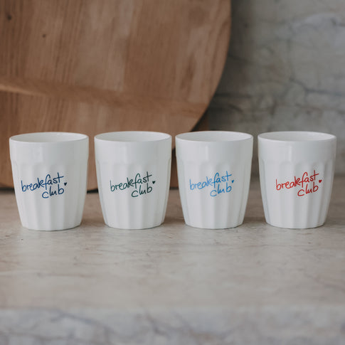 Vier weiße Keramikbecher „breakfast club“ mit farbigen Schriftzügen, minimalistisch arrangiert