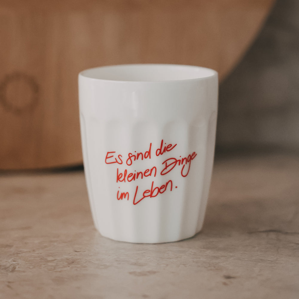 Weißer Keramikbecher mit rotem Schriftzug „Es sind die kleinen Dinge im Leben“ auf hellem Untergrund