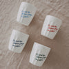 Vier weiße Becher mit Spruch „Es sind die kleinen Dinge im Leben“ in Blau, Rot und Grün auf Stoff arrangiert