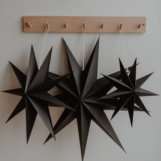 Drei schwarze Papiersterne hängen nebeneinander an einer Holzleiste mit kleinen Haken an einer weißen Wand.