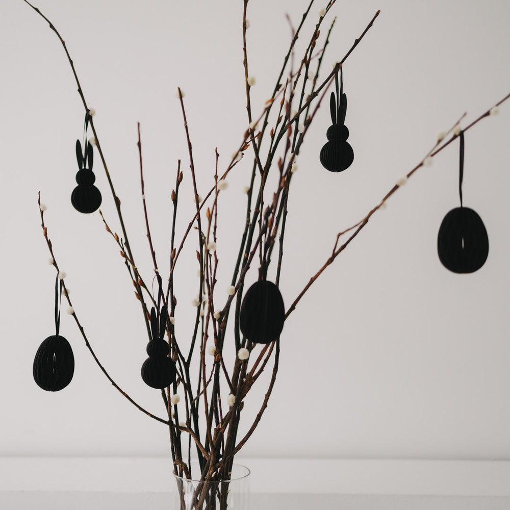 Schwarze Papier-Osteranhänger in Eiform und Hasenform an dekorierten Zweigen.