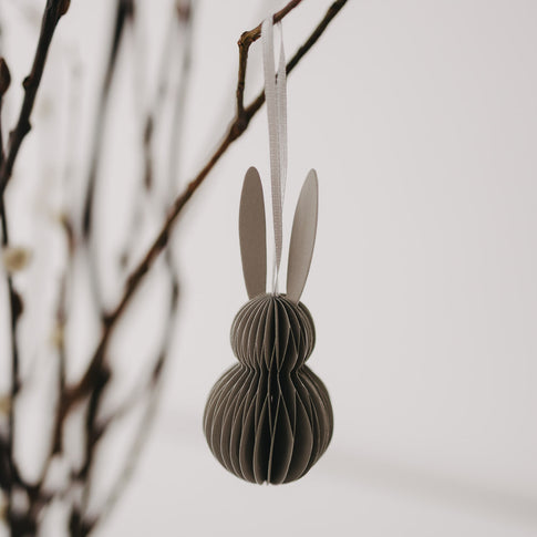 Grauer Papieranhänger in Hasenform mit weißen Ohren hängt an einem Zweig.