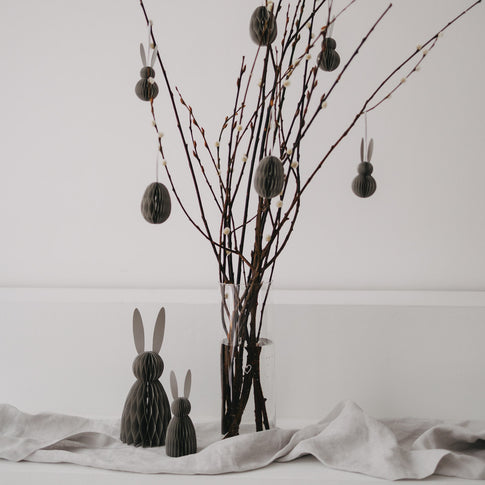 Graue Osteranhänger aus Papier hängen an Zweigen in einer Glasvase, dekoriert mit Hasenfiguren.