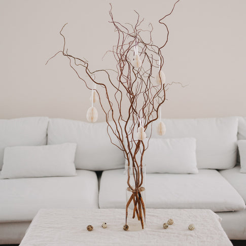 Osterstrauß mit beigen Papieranhängern in Ei- und Hasenform vor weißem Sofa.