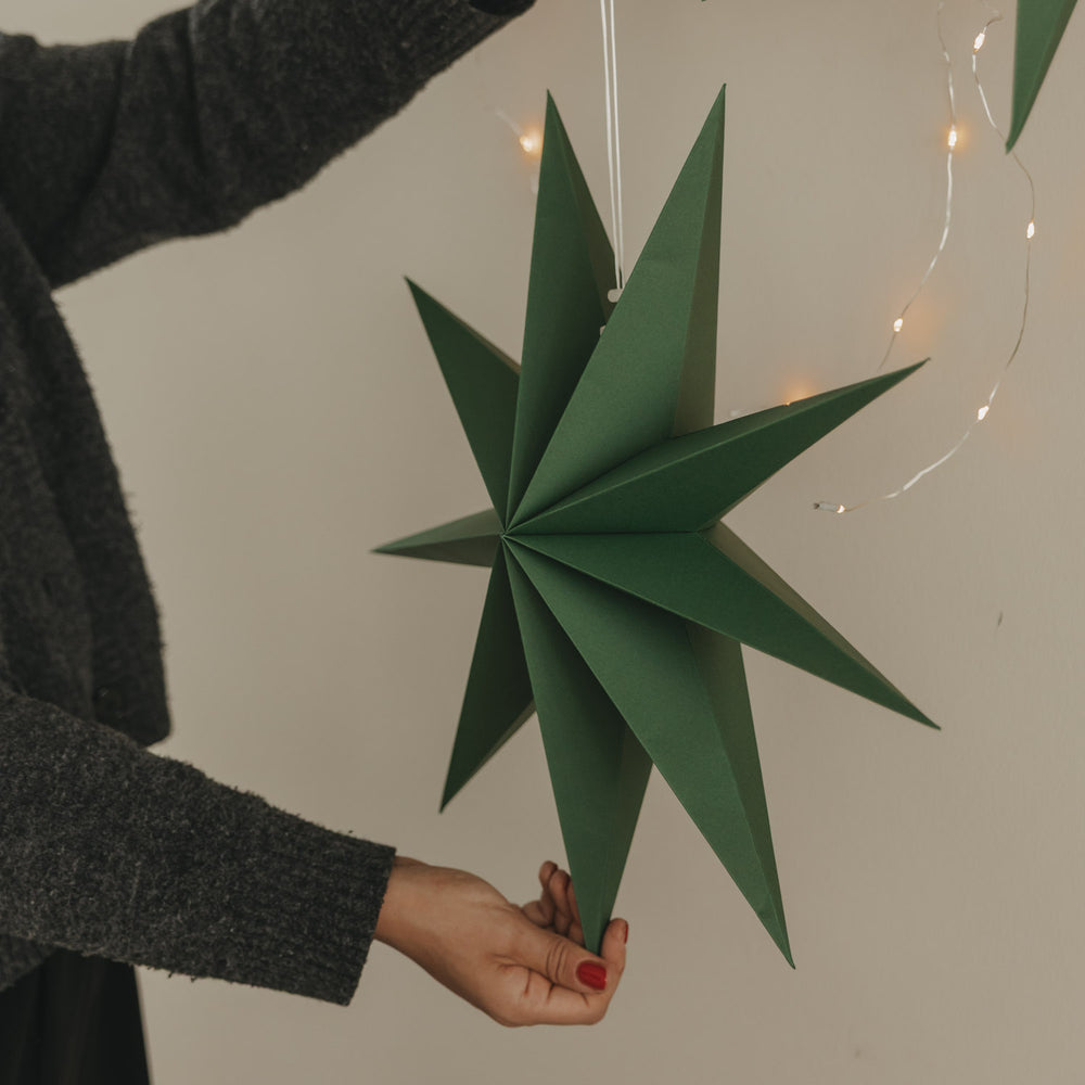 Dunkelgrüner Papierstern mit langen Spitzen und weißer Aufhängung vor warmer Lichterkette