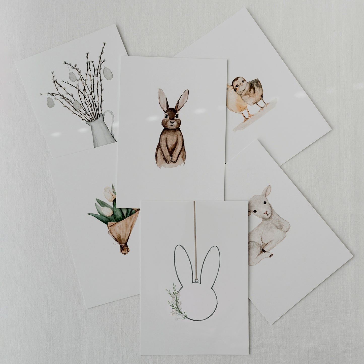 Oster-Kartenset mit sechs Aquarellmotiven locker ausgelegt: Hase, Lamm, Küken, Tulpen und Weidenzweige.