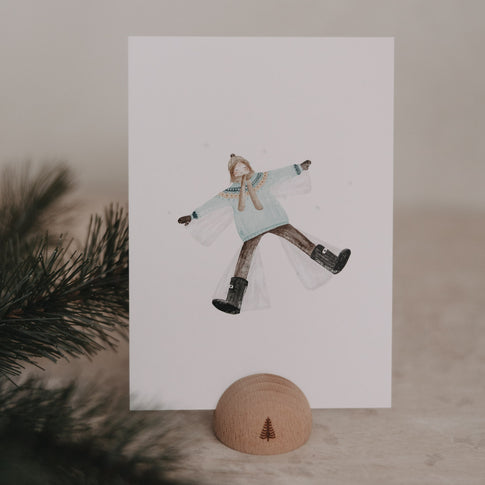 Weihnachtskarte mit Frau im Winteroutfit, die auf dem Rücken liegt und einen Schneeengel macht