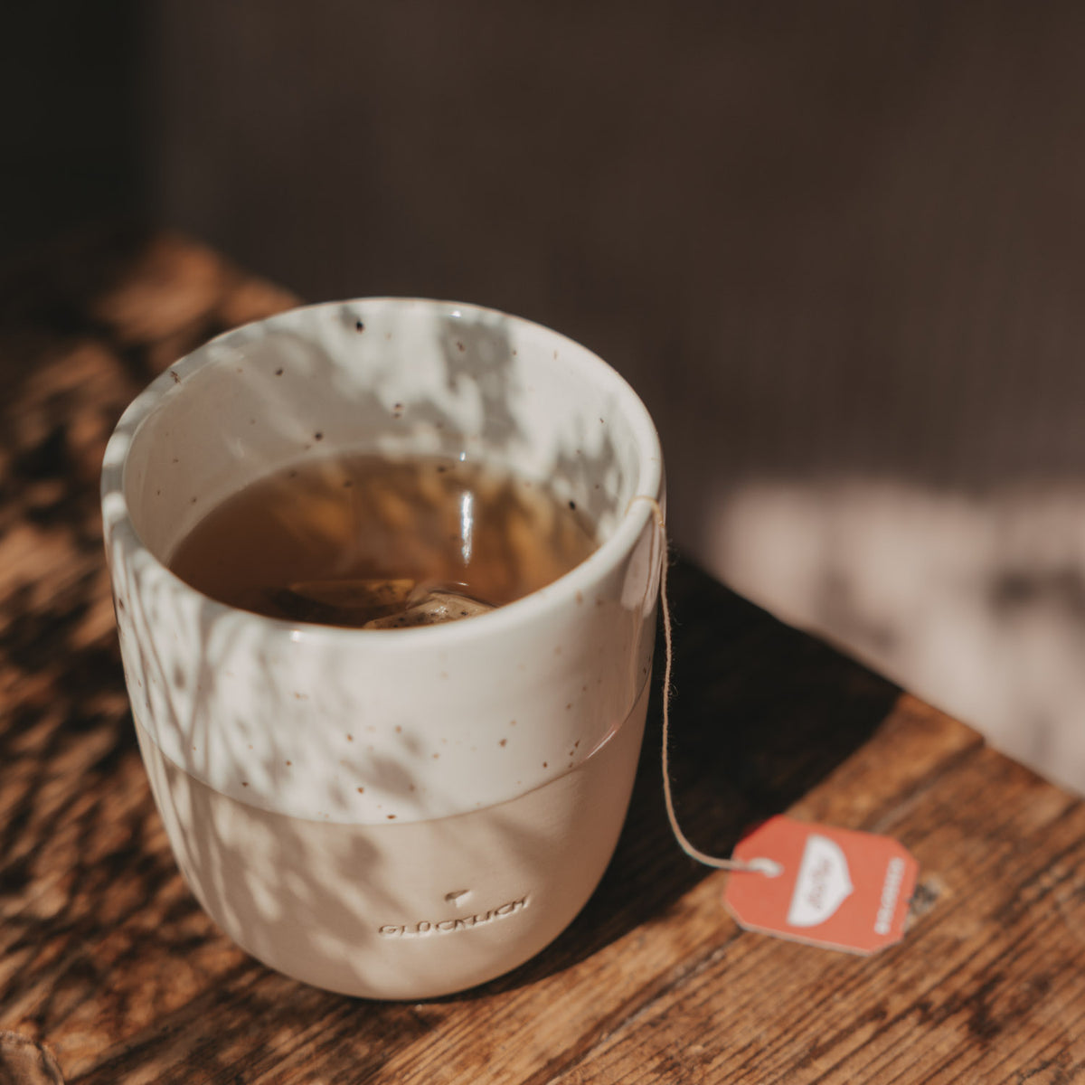 Beige-weißer Keramikbecher mit der Prägung „Glücklich“, gefüllt mit Tee und Teebeutel, im warmen Sonnenlicht auf einem Holztisch.